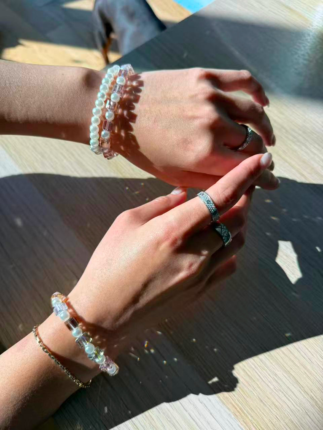 Hands stacking Aiko Tanaka Kosai no Wa pearl and crystal cube bracelets with rings on a sunlit wooden table