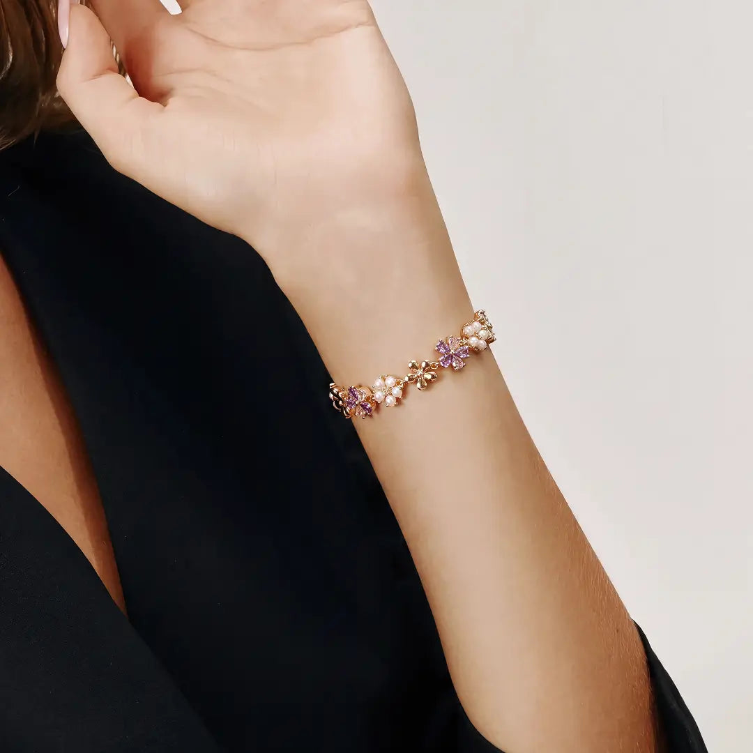 Aiko Tanaka Kasen bracelet, a gold chain with pink flowers and pearls, worn on a raised wrist against a black top