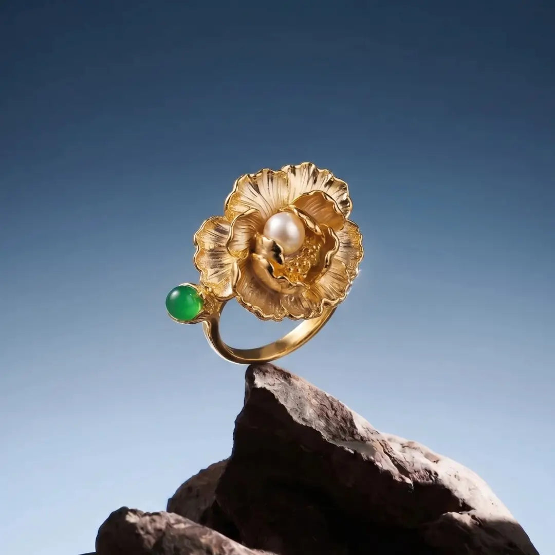 Aiko Tanaka Kinsenka ring, a gold flower ring with a pearl and green stone, displayed on dark rock against blue sky