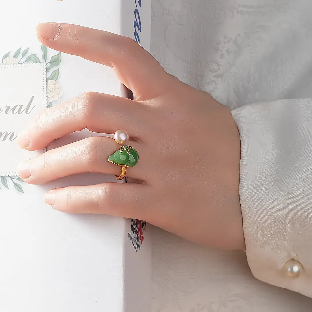 Hand in a white sleeve wearing the Aiko Tanaka Kissho ring, a green jade gourd beside a pink pearl