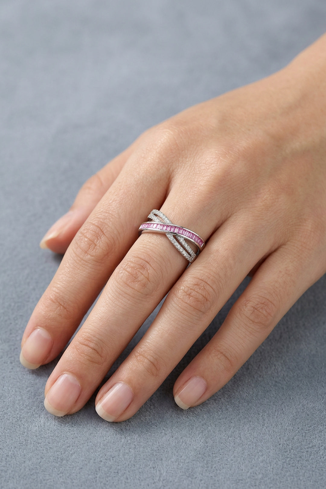 Hand resting on soft blue fabric wearing the Aiko Tanaka Kosa silver crossover ring with pink and clear stones