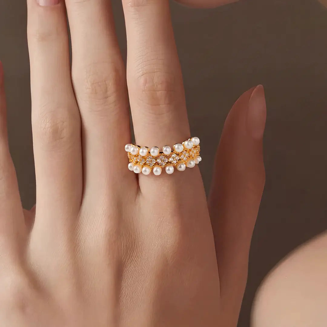 Aiko Tanaka Rinka ring, a gold band with tiers of small pearls, worn on the middle finger against a brown backdrop