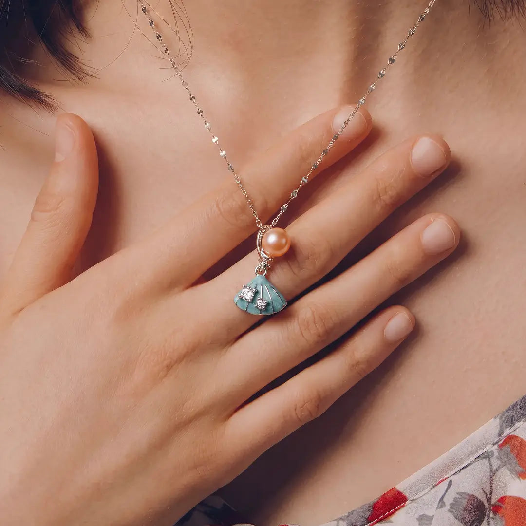 Aiko Tanaka Suiren necklace worn at the collarbone, a hand resting near the peach pearl and blue enamel pendant