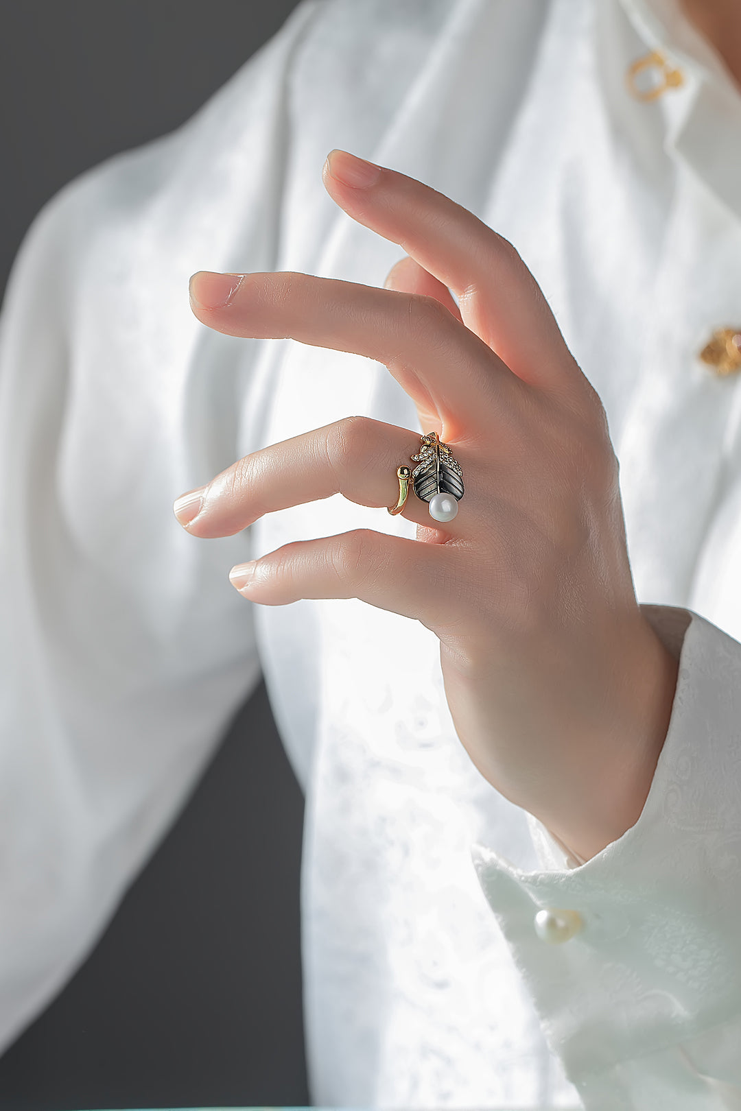 Hand in a white blouse wearing the Aiko Tanaka Yoru no Ha dark leaf and pearl ring against a grey background